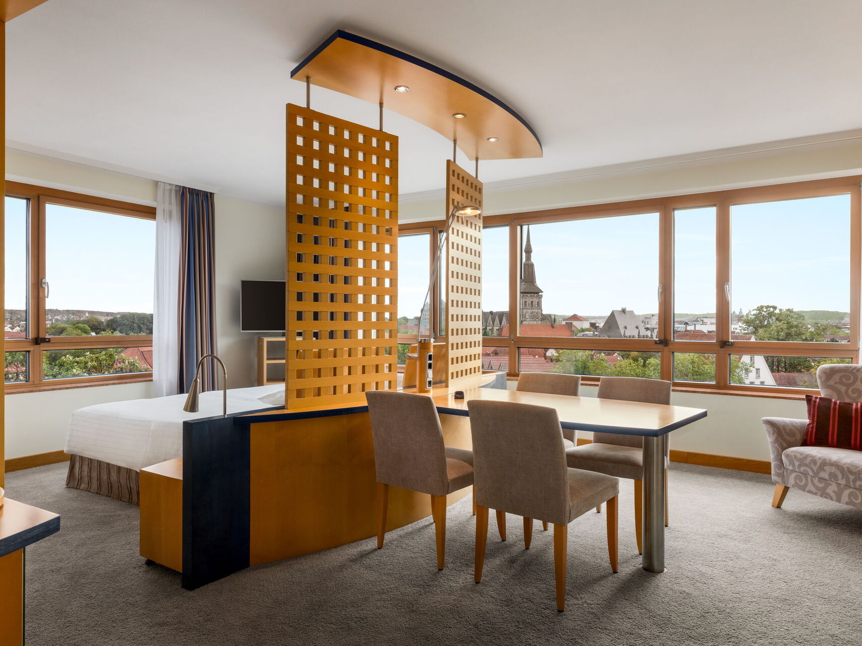 Hotel Remarque Panorama Suite mit Doppelbett, Fernseher und offenem Wohnbereich mit Tisch für 4 Personen, Stühlen, Sofa, Fensterfront mit Ausblick auf die Stadt
