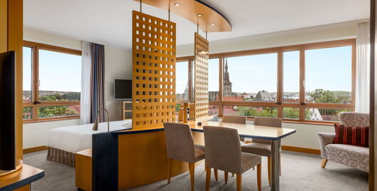 Hotel Remarque Panorama Suite mit Doppelbett, Fernseher und offenem Wohnbereich mit Tisch für 4 Personen, Stühlen, Sofa, Fensterfront mit Ausblick auf die Stadt